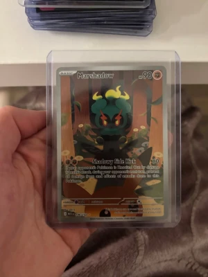 Pokémonkort Marshadow holo - Säljer ett Pokémonkort med Marshadow-motiv, holofoil och färgstark design. Kortet har en mörk bakgrund med gula och gröna detaljer och är skyddat i en plastficka. Perfekt för samlare eller Pokémonfans. Mega evolution. Potential till 10 graderad. 