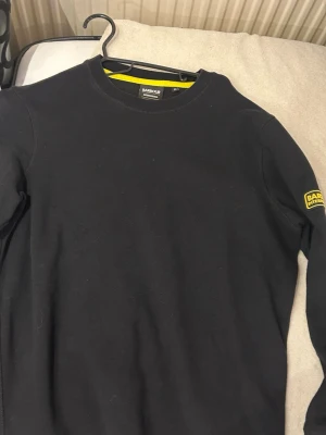 Svart sweatshirt från Barbour - Klassisk svart sweatshirt från Barbour International med rund halsringning och diskret logga på ärmen. Tröjan har en enkel och stilren design med ribbade muddar och mjukt material, perfekt för en avslappnad look.