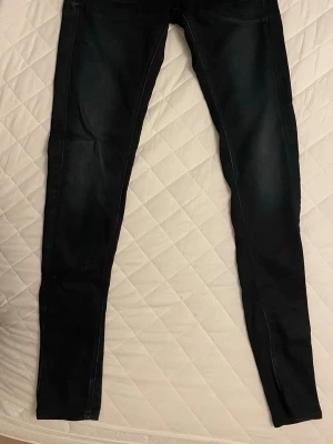 Mörkblå skinny jeans från G-Star - Säljer ett par mörkblå skinny jeans från G-Star med klassisk femficksdesign och diskreta slitningar. Jeansen har en smal passform och detaljerade sömmar på bakfickorna samt en svart patch med logga i midjan. Perfekta för en clean och stilren look.