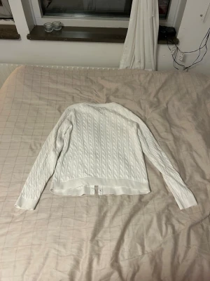 Vit kabelstickad tröja - Snygg vit kabelstickad tröja med långa ärmar och ribbade muddar. Tröjan har klassiskt mönster och är perfekt för lager-på-lager. Passar dig som gillar en stilren och enkel look.