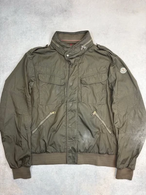 Olivgrön vindjacka från Moncler - Snygg olivgrön vindjacka från Moncler med hög krage, dragkedja och knappstängning. Jackan har två bröstfickor med lock, två snedställda fickor med dragkedja och ribbade muddar vid ärmslut och nederkant. Moncler-logga på vänster ärm. Tillverkad i lätt, vindtätt material.