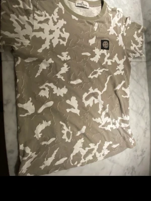 Beige camo t-shirt från Stone Island - Säljer en beige t-shirt från Stone Island med vit camouflage-mönster och klassisk logopatch på bröstet. T-shirten har rund hals och korta ärmar. Perfekt för dig som gillar streetwear och vill sticka ut lite extra.