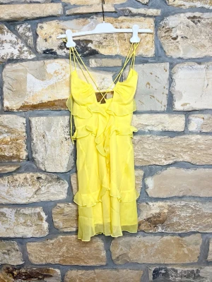 Gul volangklänning med tunna band - Söt gul kortklänning med tunna axelband och volangdetaljer framtill och längs fållen. Klänningen har en lätt och luftig känsla, perfekt för varma dagar