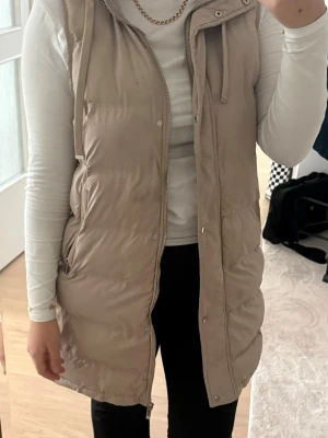 Beige lång dunväst med huva - Snygg beige lång dunväst med huva och dragkedja framtill. Västen har tryckknappar, fickor på sidorna och quiltad design. Perfekt att slänga över en hoodie eller tröja för extra värme. Passar perfekt nu till våren. Flitigt använd men är ff i fint skick.