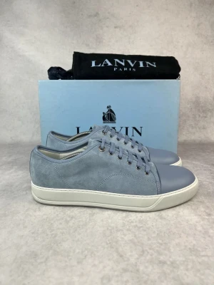 Blå sneakers från Lanvin i mocka - Vi har skorna i 41 och 43. Snygga blå sneakers från Lanvin med ovandel i mjuk mocka och tåparti i matchande blått läder. Skorna har vita sulor och klassisk låg profil med blå skosnören och metallöljetter. Perfekta för en clean och stilren look.