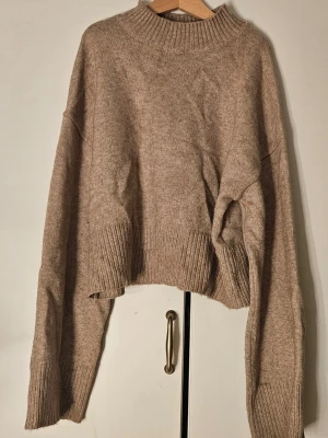 Beige stickad tröja - Mysig beige stickad tröja med croppad passform och bred ribbad mudd vid ärmslut och nederkant. Tröjan har hög hals och avslappnad siluett, perfekt för lager på lager. Materialet känns mjukt och värmande, och färgen är lätt att matcha till olika outfits. Nästan aldrig använd. 