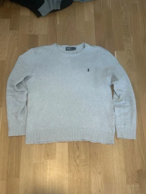 Ljusgrå stickad tröja Polo Ralph Lauren - Ljusgrå stickad tröja från Polo Ralph Lauren med rund halsringning och diskret broderad logotyp på bröstet. Tröjan har ribbade muddar vid ärmslut och nederkant. Perfekt för lager på lager och enkel att matcha.