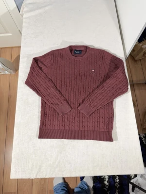 Bondelid kabelstickad tröja - Säljer en snygg vinröd kabelstickad tröja från bondelid. Tröjan har rund halsringning, ribbade muddar och nederkant samt en liten bondelid-logga broderad på bröstet. Tröjan är i fint sckik.