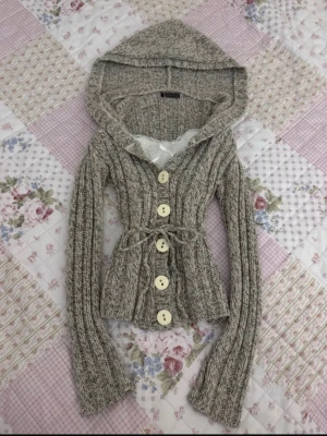 Gråmelerad stickad kofta med huva - Mysig gråmelerad stickad kofta med stora vita knappar och huva. Koftan har ett knytband i midjan och ribbstickat mönster. Perfekt för lager-på-lager och chill dagar. Långa ärmar och figurnära passform.