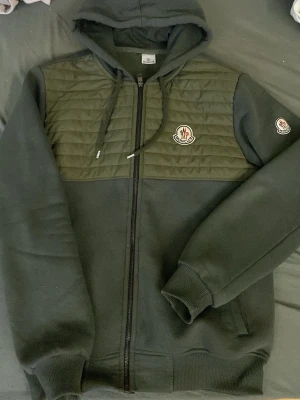 Mörkgrön Moncler hoodie med dragkedja - Pris kan diskuteras vid snabb affär..Snygg mörkgrön hoodie från Moncler med dragkedja framtill och quiltad panel över bröstet. Har huva med dragsko, ribbade muddar och två fickor. Moncler-logga på bröstet och ärmen. Perfekt för en chill och trendig look.