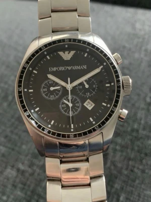 Emporio Armani silverfärgad klocka - Stilren klocka från Emporio Armani i silverfärgat rostfritt stål med svart urtavla. Klockan har kronograf, datumvisning och länkarmband. Klassisk design med tydliga index och logga upptill på urtavlan.