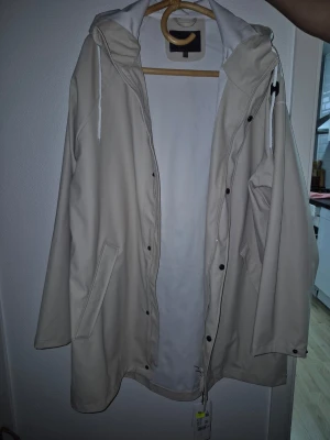 Beige regnjacka med huva - Stilren beige regnjacka med huva och dragsko, knäppning med tryckknappar och två snedställda fickor framtill. Jackan har en rak passform och är tillverkad i ett vattenavvisande material, perfekt för regniga dagar.