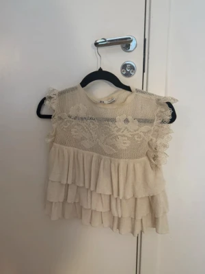 Beige volangblus med spetsdetaljer - Supersöt beige blus med volanger och spets upptill. Ärmlös modell med romantiska detaljer och rund halsringning. Perfekt för dig som gillar en feminin och trendig stil.