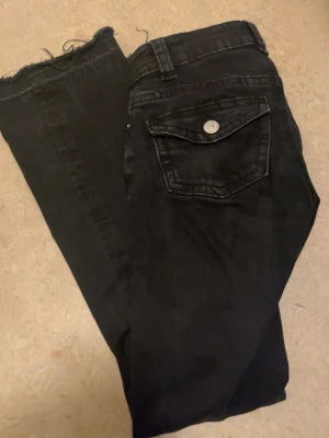 Svarta jeans med rå kant - Säljer ett par svarta jeans med rå, avklippt kant nedtill och cool fickdetalj med knapp bak. Jeansen har en klassisk femficksmodell och är i ett mjukt denimtyg. Perfekta för en avslappnad och trendig look. Lågmidjade 