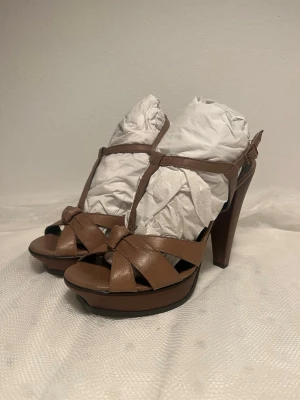 Rizzo Bruna platåpumps med korsade remmar - Från Rizzo.Snygga bruna pumps med hög platåklack och korsade remmar framtill. Skorna har öppen tå och T-rem över foten för extra stöd. Tillverkade i slätt läder med en elegant siluett som ger höjd och stil till din outfit.