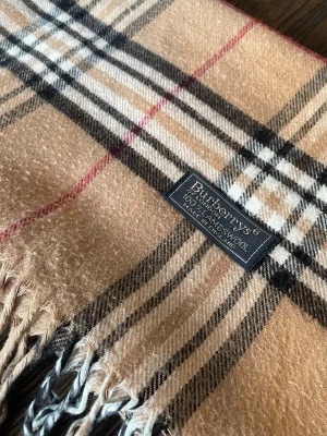 Beige rutig halsduk från Burberry - Klassisk beige halsduk från Burberry i 100% lammull med det ikoniska rutmönstret i svart, vitt och rött. Halsduken har fransar i ändarna och en mjuk, lyxig känsla. Perfekt accessoar för att lyfta din outfit med en tidlös brittisk stil.
