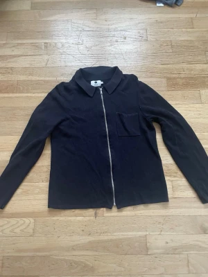 Svart ziptröja från NN07 - Stilren svart ziptröja från NN07 med krage och bröstficka. Tröjan har långa ärmar och är tillverkad i ett mjukt, stickat material. Perfekt för en clean och avslappnad look.