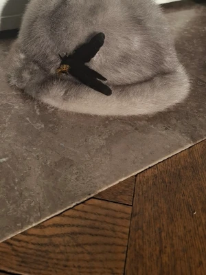 Grå fluffig hatt  - Säljer en supermjuk och fluffig grå hatt med en svart dekorativ fjädrar på sidan. Hatten har rund form och bred kant, perfekt för dig som vill sticka ut med en unik accessoar.