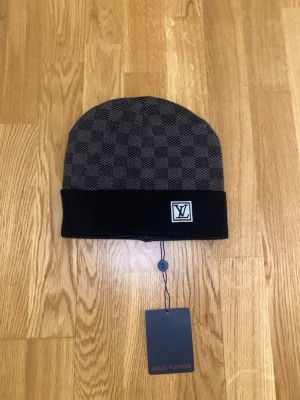 Mörkgrå mössa - Snygg mörkgrå mössa med klassiskt rutmönster och vikt kant. Mössan har en diskret logga framtill och är tillverkad i mjukt, stickat material som känns lyxigt och bekvämt.