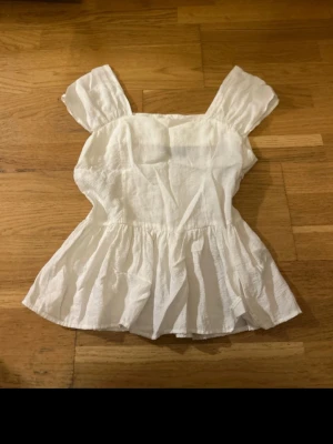 Vit volang topp med breda axelband - Söt vit topp med breda axelband och volang nedtill. Tillverkad i ett lätt och luftigt tyg, perfekt för varma dagar. Toppen har en feminin siluett och är ärmlös, vilket ger en fräsch och somrig känsla.