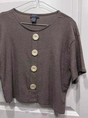 Brun topp med stora knappar från Primark - Kortärmad brun topp från Primark med fyra stora dekorativa knappar framtill. Toppen har en rund halsringning och är croppad i modellen. Materialet känns mjukt och lätt, perfekt för en chill och stilren look.