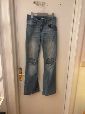 Ljusblå slitna bootcut jeans - Säljer ett par ljusblå bootcut jeans med slitningar och hål på knäna. Jeansen har klassisk femficksdesign, låg midja och är tillverkade i denim. Perfekta för en avslappnad och trendig look.