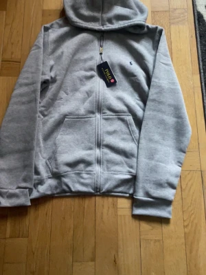 Grå zip-hoodie från Polo Ralph Lauren - Snygg grå hoodie med dragkedja från Polo Ralph Lauren. Klassisk design med huva, två fickor framtill och liten broderad logga på bröstet. Mjuk bomullsblandning som är perfekt för en avslappnad stil.