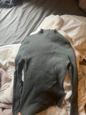 Grå/grön ribbad polotröja från H&M - Säljer en stilren grå ribbstickad polotröja från H&M. Tröjan har lång ärm, hög krage och är tillverkad i ett mjukt, stretchigt material som sitter snyggt på kroppen. Perfekt för lager på lager och enkel att matcha med olika outfits.