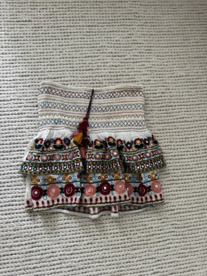 Broderad kjol med volanger - Unik skort med broderade blommor i flera färger, volangkant och smockad midja. Basen är vit med detaljer i rött, blått, gult och grönt samt dekorativ knytning med tofsar framtill. Perfekt för dig som gillar boho-stil och färgglada plagg.