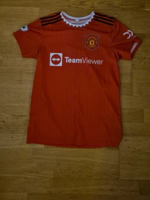 Manchester United matchtröja Elanga - Sporttröja av Manchester United matchtröja  med Elanga och nummer 36 på ryggen. Tröjan har vita detaljer vid kragen, svarta ränder på axlarna och Premier League-logga på ärmen. Materialet är lätt och andas, perfekt för fotbollsträning eller match. GÅR ATT PRUTA