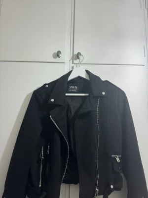 Svart mockajacka från ZARA - Snygg svart bikerjacka från ZARA med silvriga dragkedjor och knappar. Jackan har flera fickor med dragkedja och detaljer som ger en cool look. Tillverkad i mjukt material (locka) som känns bekvämt. Jackan har använts få gånger och kan bäras i storlekarna Xss-S