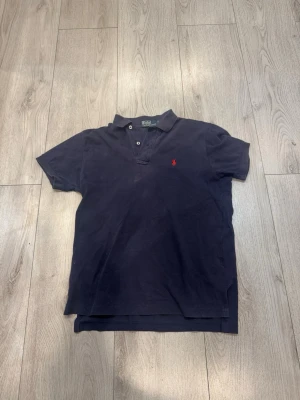 Mörkblå pikétröja från Ralph Lauren - Klassisk mörkblå pikétröja från Ralph Lauren med röd broderad logga på bröstet. Tröjan har korta ärmar, krage och knappar vid halsen. Tillverkad i mjuk bomull som känns skön mot huden. Perfekt till jeans eller shorts för en avslappnad look. Storlek S men sitter som M