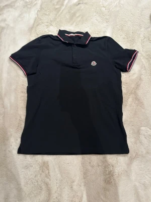 Riktigt snygg Svart pikétröja med rödvit kant - Klassisk svart pikétröja med krage och knappar framtill. Tröjan har röda och vita detaljer på krage och ärmslut samt Perfekt för en stilren och sportig look.