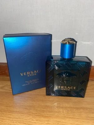 Versace Eros Eau de Toilette 100ml - Versace Eros Eau de Toilette i en elegant, fyrkantig blå glasflaska med detaljerat mönster och Versace-logga. Flaskan har ett matchande blått lock och kommer i en lyxig blå kartong med diskret mönster och silvertext. En ikonisk doft för dig som gillar exklusiva parfymer.