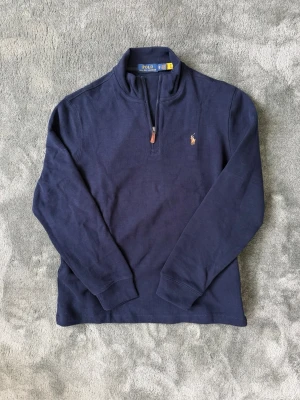 Mörkblå halvzip från Polo Ralph Lauren - Klassisk mörkblå långärmad tröja från Polo Ralph Lauren med halv dragkedja och broderad logga på bröstet. Tröjan har hög krage och är tillverkad i mjuk bomull. Perfekt för en stilren och avslappnad look. Storlek XS men passar även någon som använder S