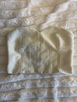 Champagnefärgad tubtopp med struktur - Snygg axelbandslös korsett topp i champagnefärgat tyg med strukturerad yta. Toppen har markerade sömmar framtill och dragkedja bak för enkel påklädning. Perfekt för att skapa en trendig look.❤️