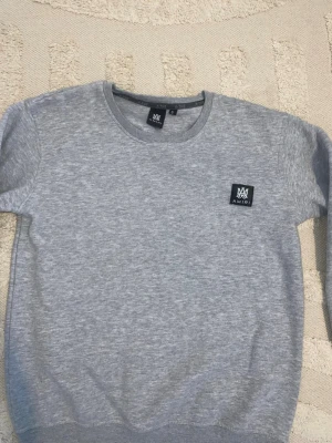 Grå sweatshirt från Amiri - Säljer en stilren grå sweatshirt från Amiri med rund halsringning och svart patch med logga på bröstet. Tröjan har ribbade muddar vid ärmslut och nederkant. Perfekt för en avslappnad och trendig look.