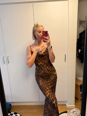 Leopardmönstrad långklänning med smala band - Snygg långklänning med leopardmönster och smala axelband. Klänningen har en figurnära passform och öppen rygg med knyt. Perfekt för dig som vill sticka ut med en djurisk look.