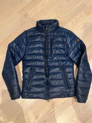 Mörkblå pufferjacka från Save the Duck  - Snygg mörkblå pufferjacka från SAVE THE DUCK med quiltad design och hög krage. Jackan har avtagbar huva, tre dragkedjeförsedda fickor och diskret logga på ärmen. Perfekt för kyliga dagar och enkel att styla till olika outfits. Mindre skada på vänster arm som knappt syns, annars hel
