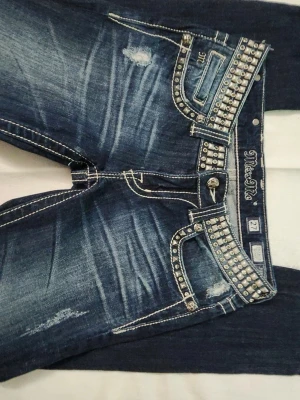 Miss Me blå jeans med nitar - Säljer ett par blå Miss Me jeans med slitningar och coola detaljer. Jeansen har vita kontrastsömmar och massor av silverfärgade nitar på fickor och linning. Materialet är denim med stretch och passformen är rak. Perfekta för dig som gillar statement-jeans!