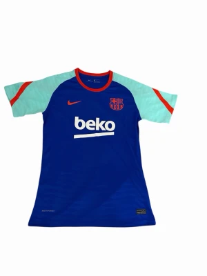 Nike FC Barcelona träningströja blå - Snygg blå FC Barcelona träningströja från Nike med röda detaljer och ljusblå ärmar. Tröjan har klubbmärke och Nike-logga på bröstet samt BEKO-tryck framtill. Tillverkad i lätt och ventilerande material, perfekt för träning eller match.