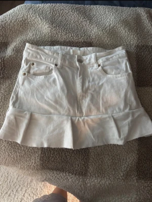 Vit jeanskjol med volangkant - Säljer en vit kort jeanskjol med volangkant nedtill. Perfekt för dig som gillar en enkel men ändå söt stil. Storlek M/38 men skulle säga att den sitter som en 34/36