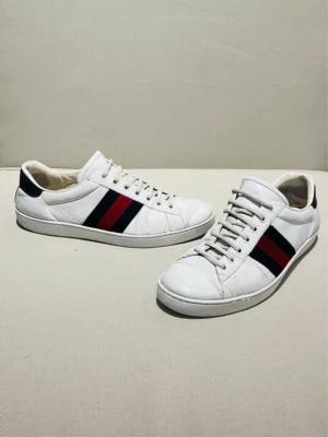 Vita Gucci sneakers med röd-blå rand - Snygga vita sneakers från Gucci med klassiska röd och marinblå ränder på sidan. Skorna har lågt skaft, vita snören och detaljer i svart på hälen. Tillverkade i läder med mönstrad yttersula. Perfekta för dig som vill ha en clean och lyxig look.
