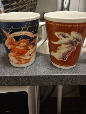 Två muggar med änglamotiv - Säljer två porslinsmuggar med juliga änglamotiv. Ena muggen är blå med texten 'Wishing you a Merry Christmas' och bild på två änglar, den andra är röd med en flygande ängel och engelsk text. Perfekta för varma drycker under vintern.