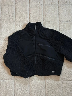 Svart teddyjacka - Säljer en svart teddyjacka med dragkedja framtill, hög krage och tre praktiska fickor med dragkedja. Jackan har en cool mix av mjukt teddy-material och släta paneler, perfekt för dig som gillar streetwear och vill ha något både snyggt och bekvämt.