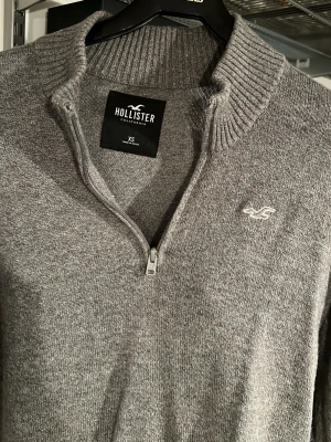 Grå stickad tröja från Hollister - Snygg grå stickad tröja från Hollister med halv dragkedja och ribbad krage. Tröjan har lång ärm och diskret Hollister-logga broderad på bröstet. Perfekt till lager-på-lager och enkel att matcha med jeans eller kjol.