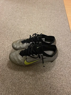 Nike Mercurial elite anniversary XXV - Snygga Nike Mercurial fotbollsskor i silver med svart och neongul swoosh. Skorna har låg profil, snörning och ovandel i syntetmaterial för bra bollkontroll. Perfekta för dig som vill ha stilrena och snabba skor på planen. Det är Elite skor och det var anniversary paret som släpptes i och med att Nike mercurial fyllde 25 (XXV) år. Väldigt sällsynta och dyra