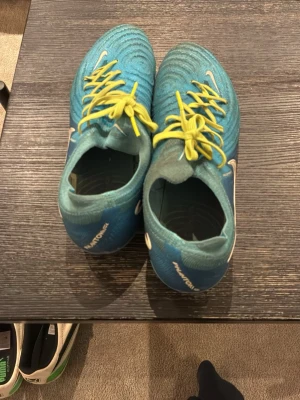 Nike Phantom blå fotbollsskor - Säljer ett par Nike Phantom fotbollsskor i blått med gula skosnören och vita detaljer. Skorna har en strumpliknande passform och är designade för bra bollkontroll på planen. Perfekta för dig som vill sticka ut med både stil och funktion. Dessa är elites så org priset ligger runt 2500-3000kr, köpte dom förra vintern och användes fram till sommaren, bytte snören då jag råkade ta sönder ena… 
