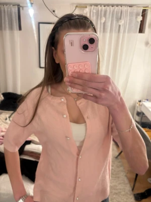 Ljusrosa kofta med pärlknappar - Söt ljusrosa kofta med korta ärmar och pärlknappar framtill.och är stickad i ett mjukt material. Perfekt att slänga över ett linne för en chill look.