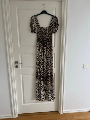 Långklänning med leopardmönster - Snygg långklänning med leopardmönster i beige, brunt och svart. Klänningen har korta puffärmar och en smockad överdel med fyrkantig ringning. Perfekt för dig som vill sticka ut med ett djurmönstrat plagg.